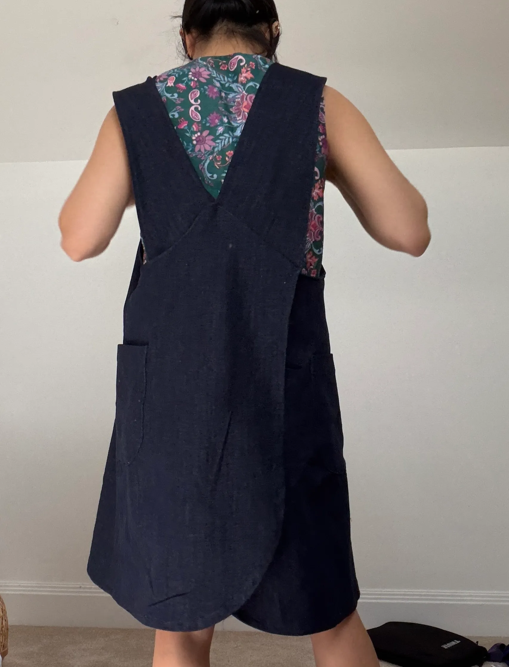 Back view of the pinafore showing the crossed shoulder straps over a green floral top, with patch pockets and a curved hem