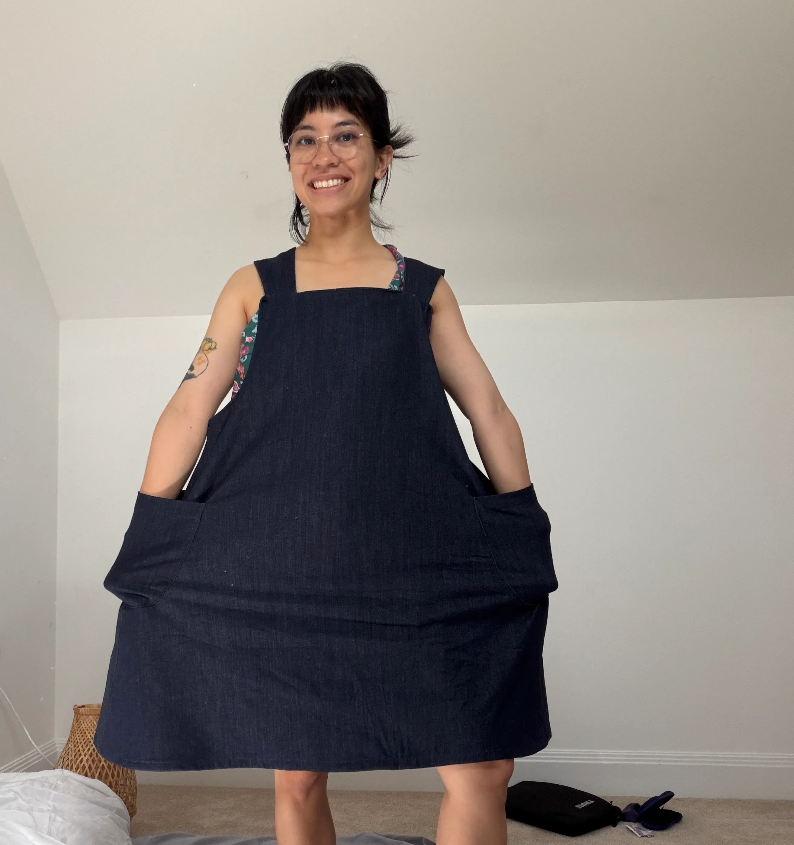 Celina smiling, holding out the two large front patch pockets of the navy denim pinafor